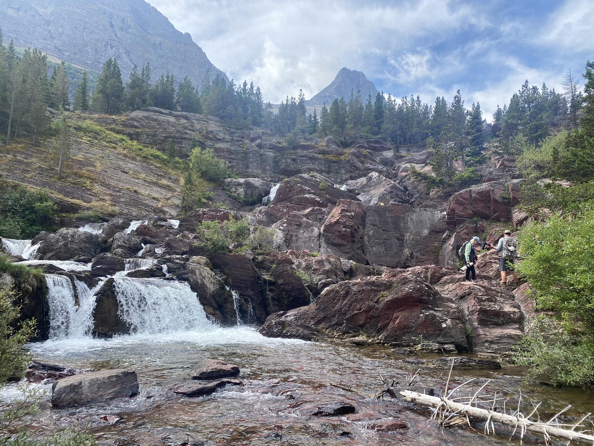 Two hikers navigate rocky terrain beside a cascading waterfall under a cloudy sky.