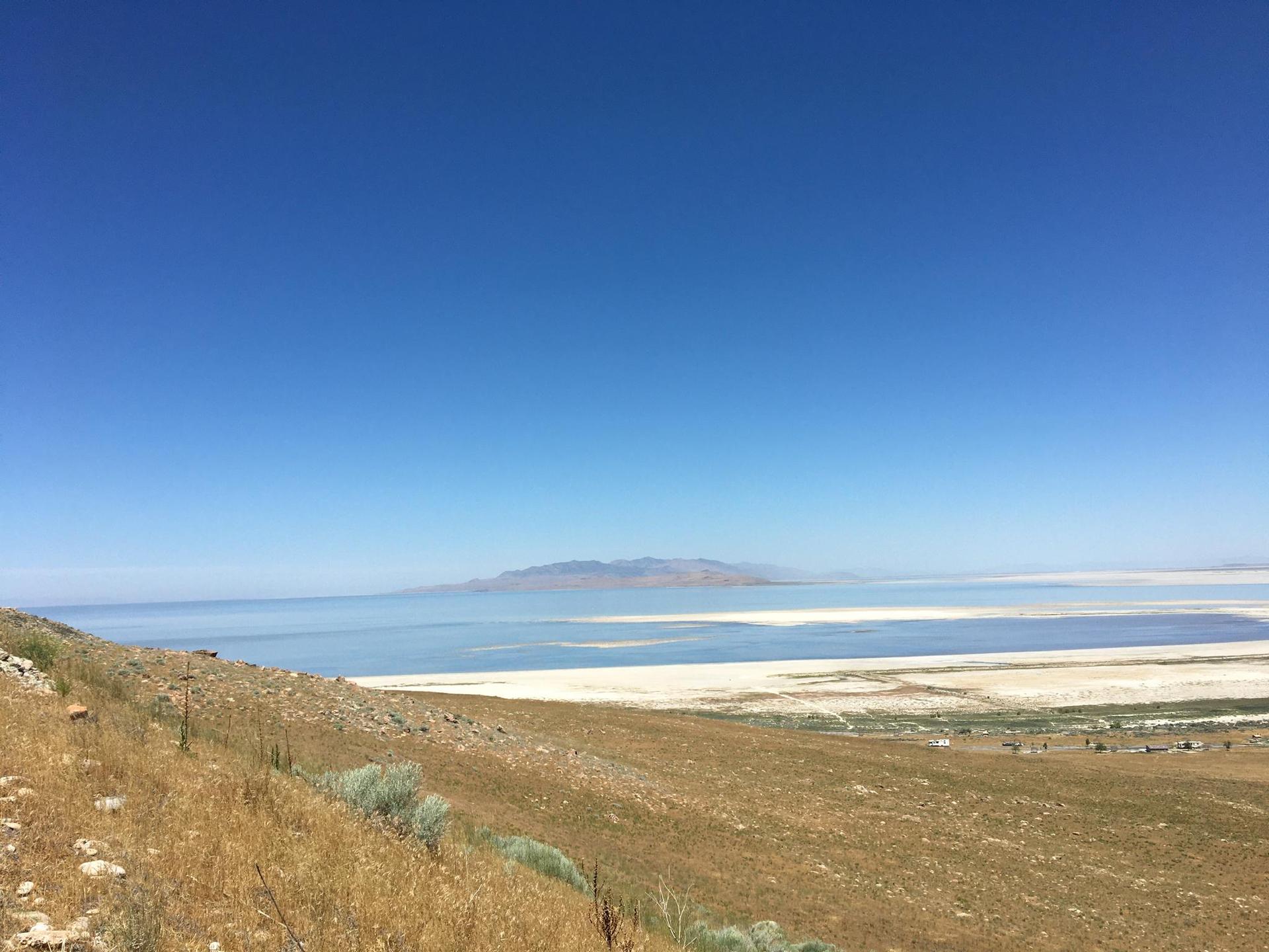 Sunny view of a vast lake, distant land, and dry, grassy hills.