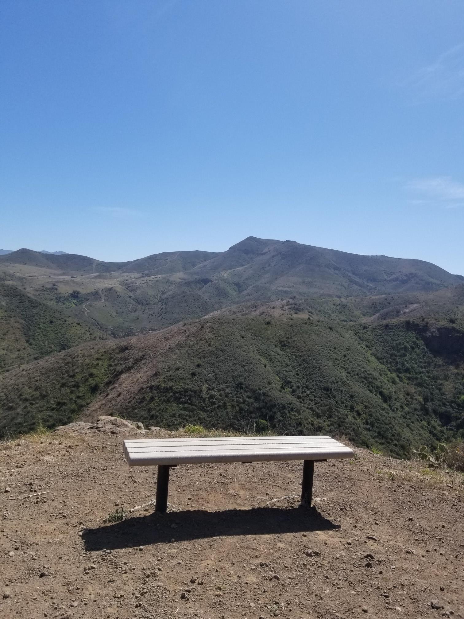 A lone bench on a hilltop overlooking vast, sunny mountain ranges.