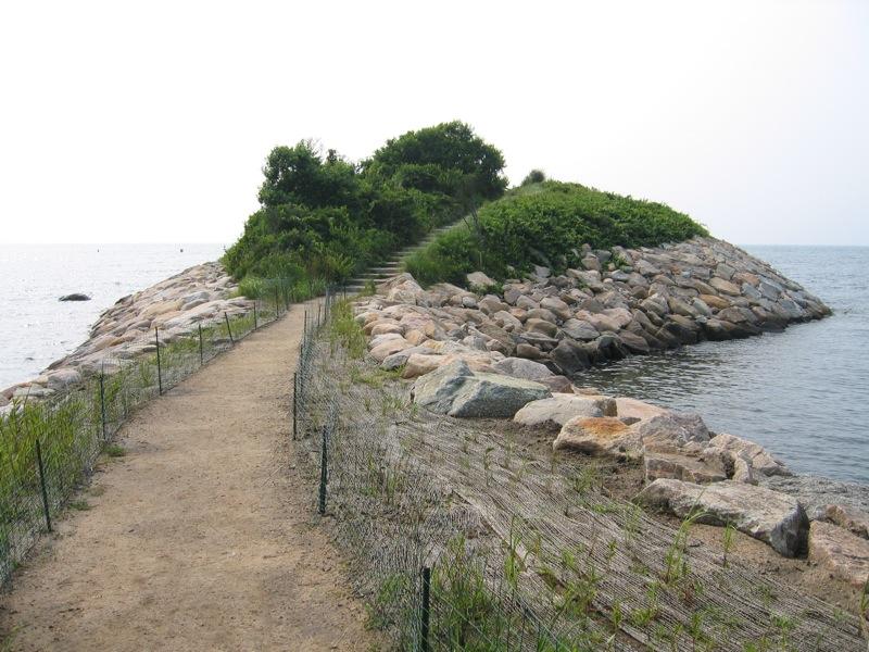 A fenced dirt path leads to a small, rocky, tree-covered island.