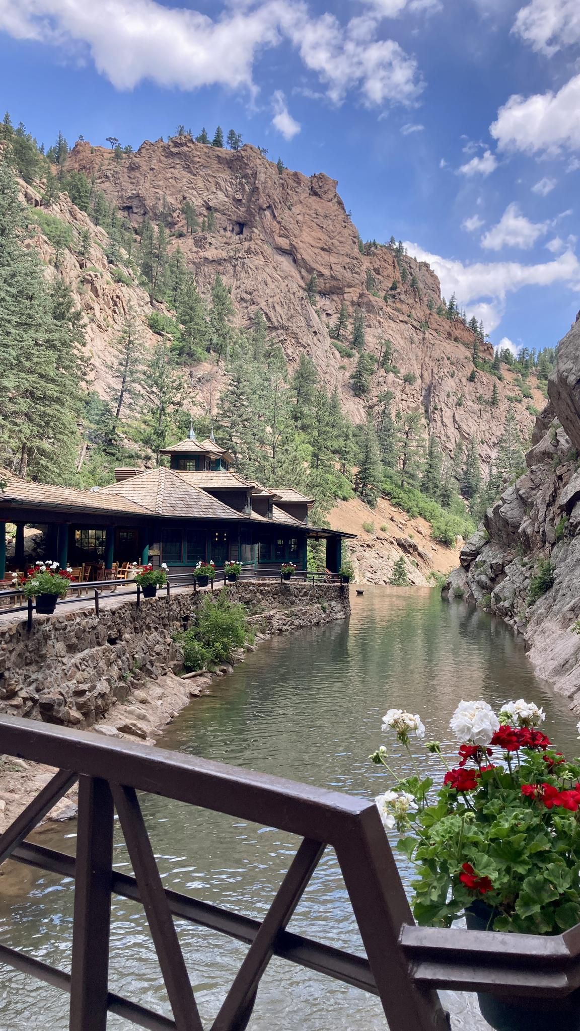 A building sits by a calm river, surrounded by steep, rocky mountains under blue sky. - Seven Falls loop from The Broadmoor