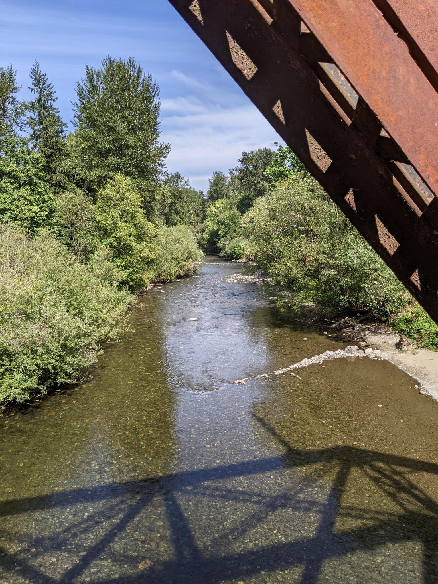 A clear river flows under a rusty metal bridge on a sunny day.