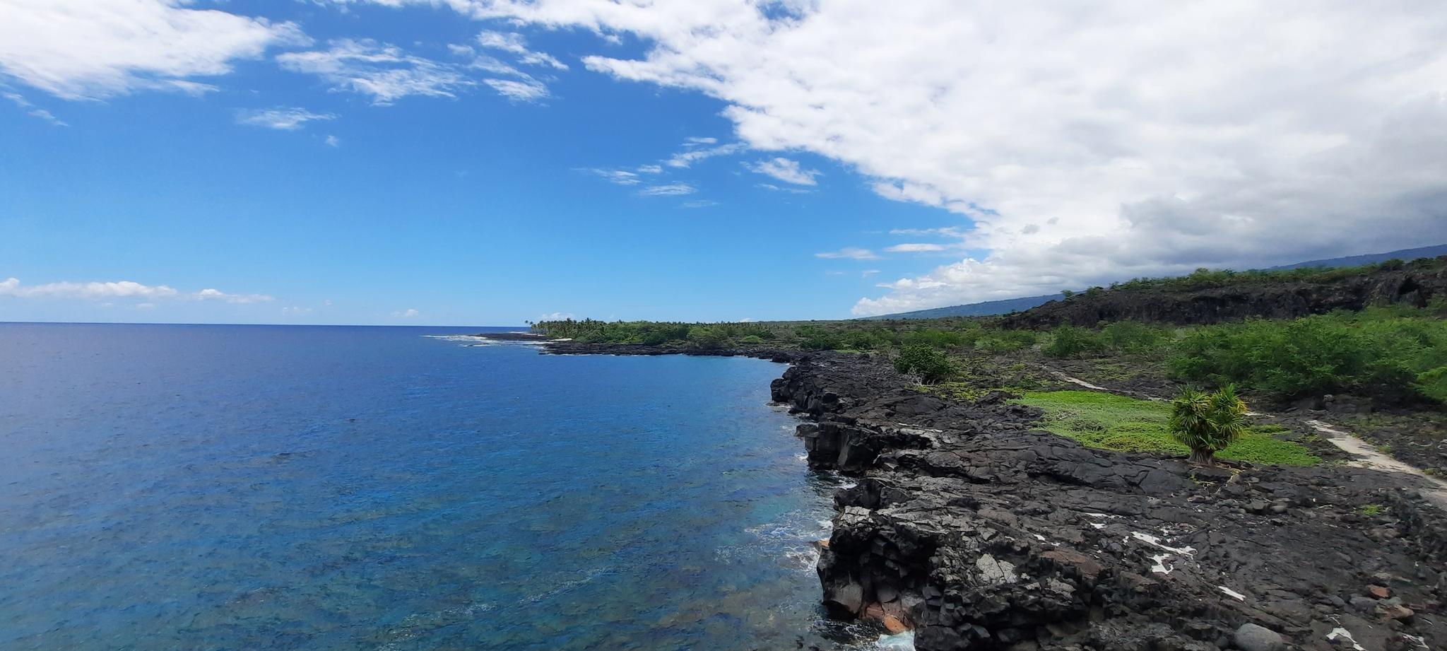 Blue ocean meets a rocky coastline with green foliage under a partly cloudy sky.