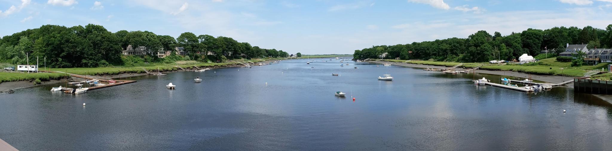 Wide river with anchored boats, flanked by green trees and houses under a bright sky.