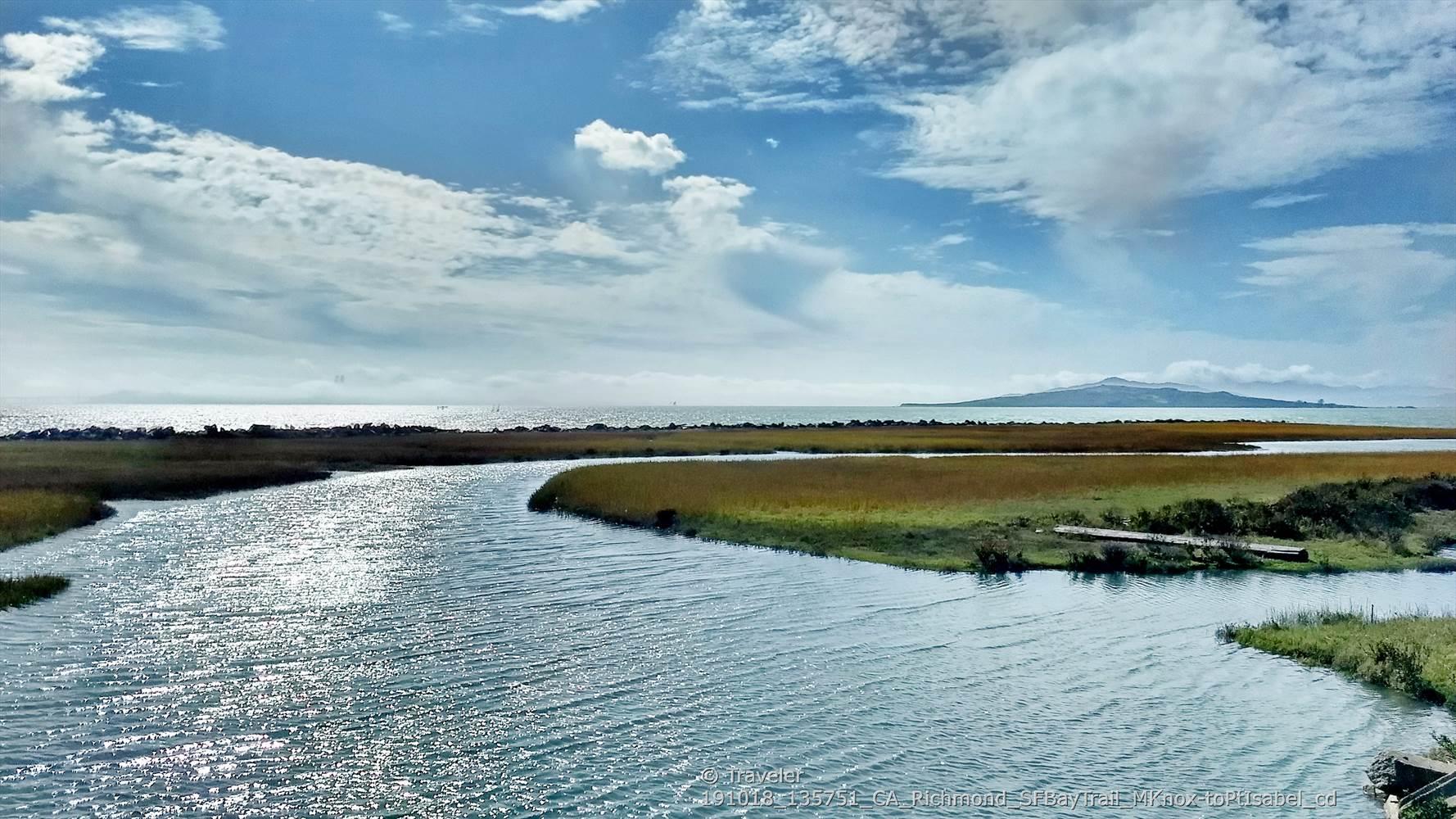 Winding river through marshland under a bright, cloudy sky, with distant ocean.
