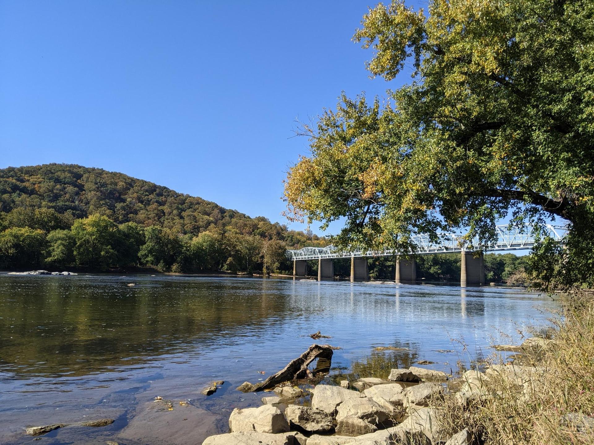 River with a distant bridge, wooded hills, and a clear blue sky.