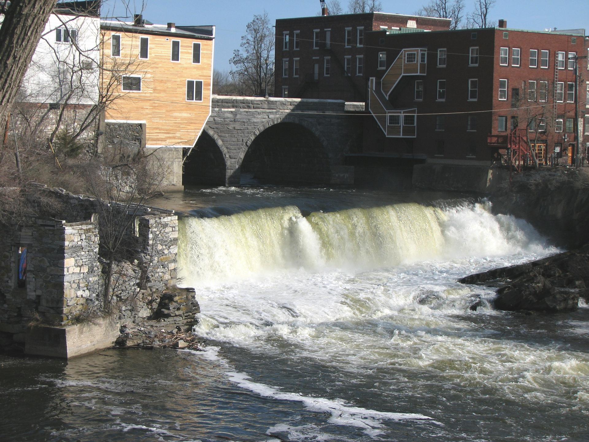 Powerful waterfall flowing under a stone arch bridge with old buildings nearby.