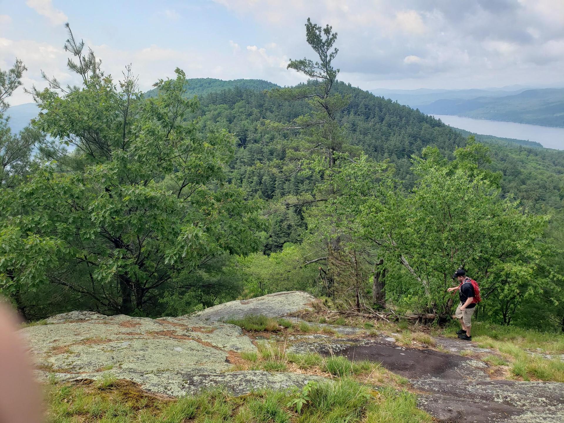 Person on rocky overlook with forested mountains and distant water under bright sky.