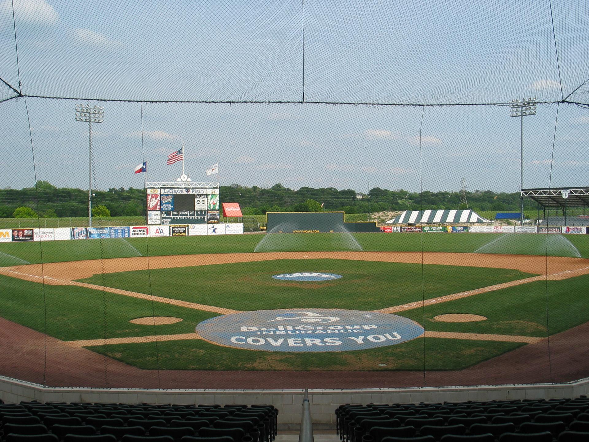 Empty baseball field with green grass and dirt bases under a cloudy sky.