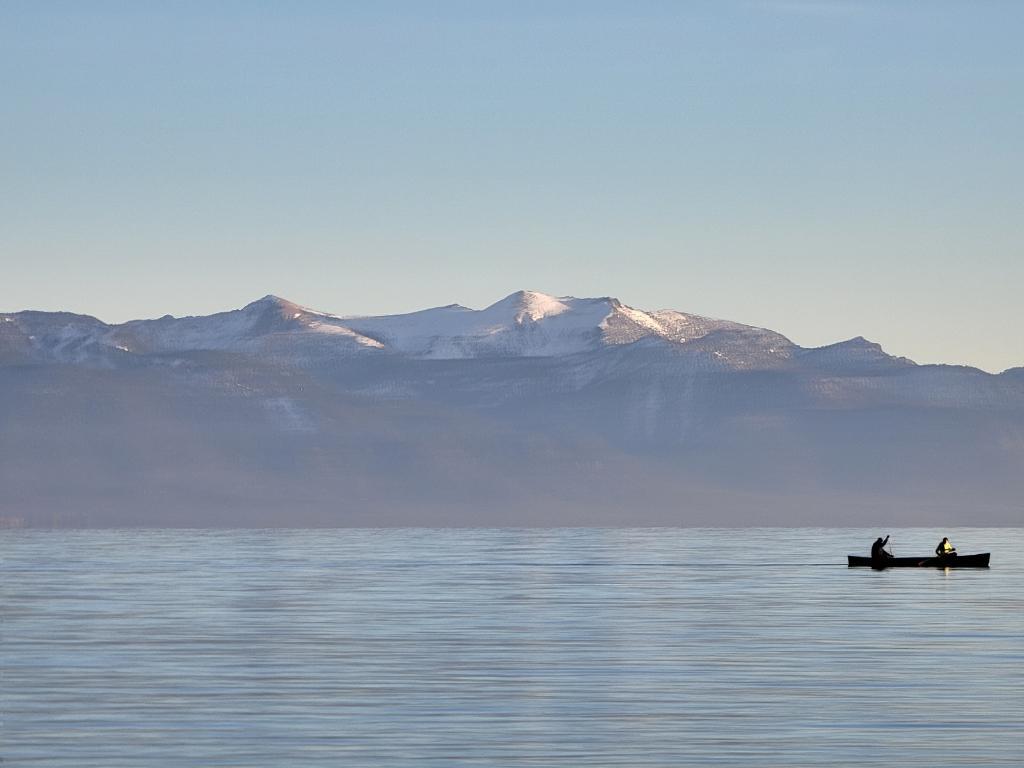 Two people canoeing on calm water with snow-capped mountains in background.