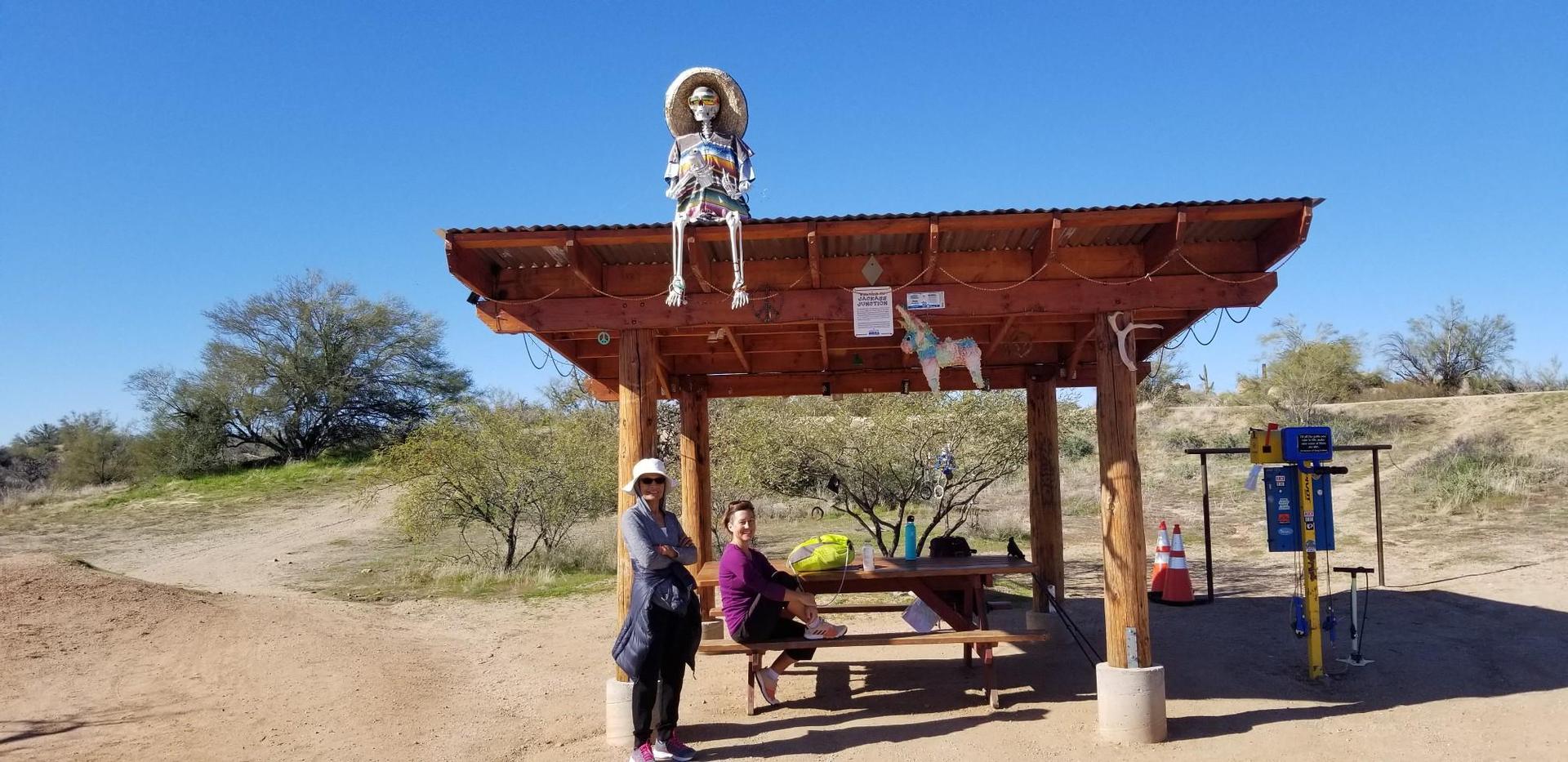 Two people relax under a wooden shelter with a skeleton figure on a sunny day.