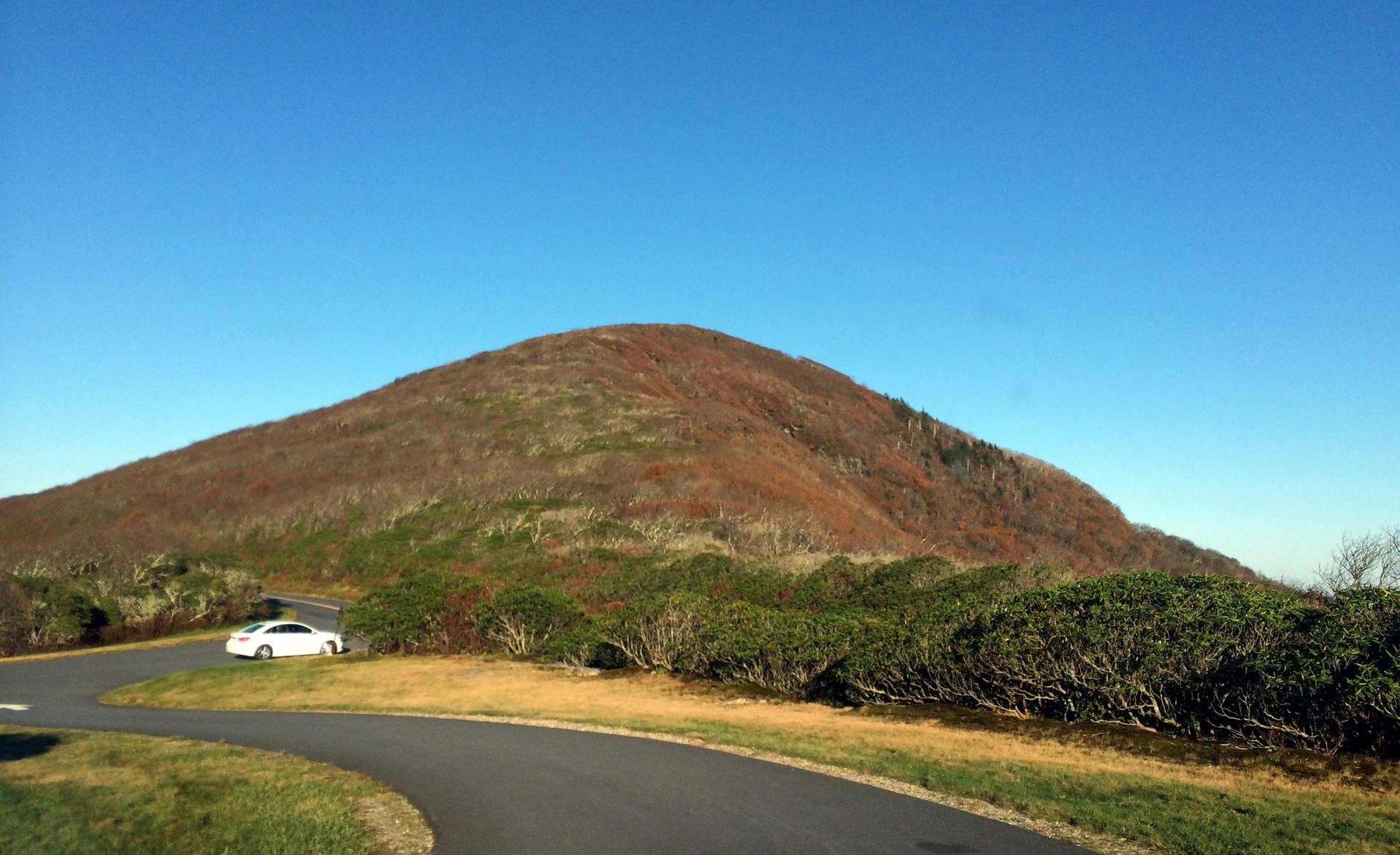 A white car parked on a winding road next to a large, reddish hill.