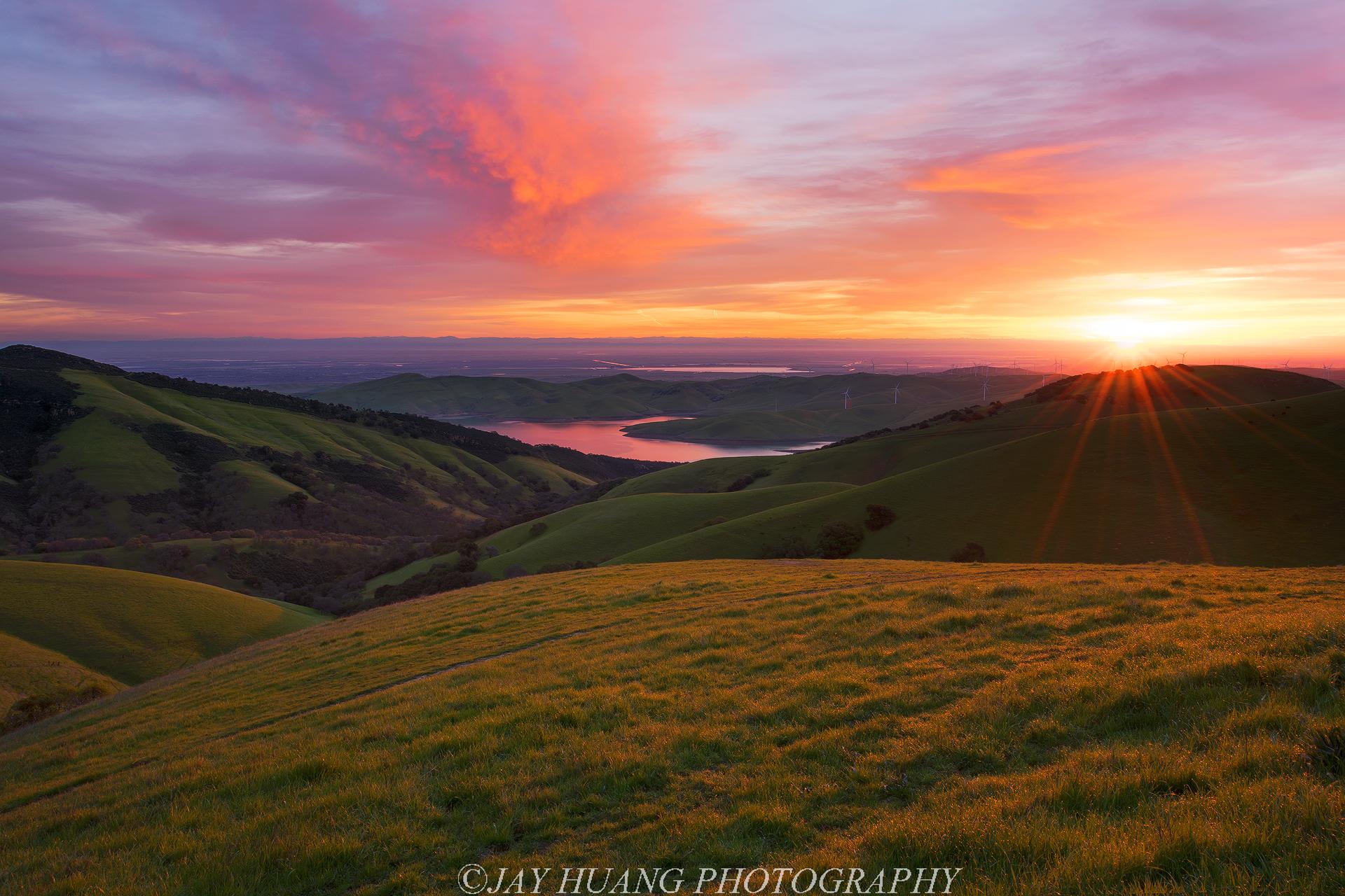 Vibrant sunset over rolling green hills, a valley, and distant water.