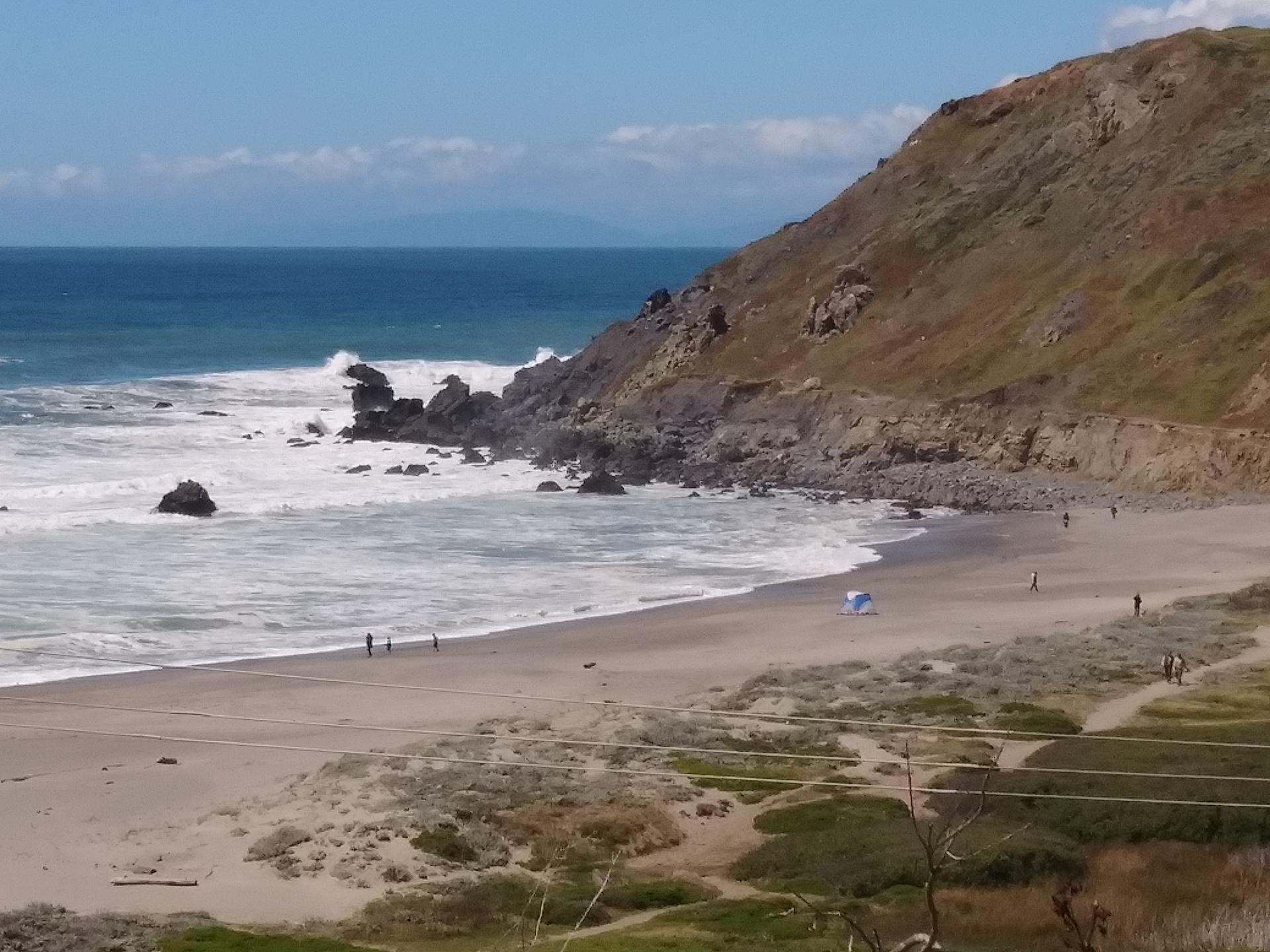 Sunny beach with crashing waves, rocky cliffs, and distant people.