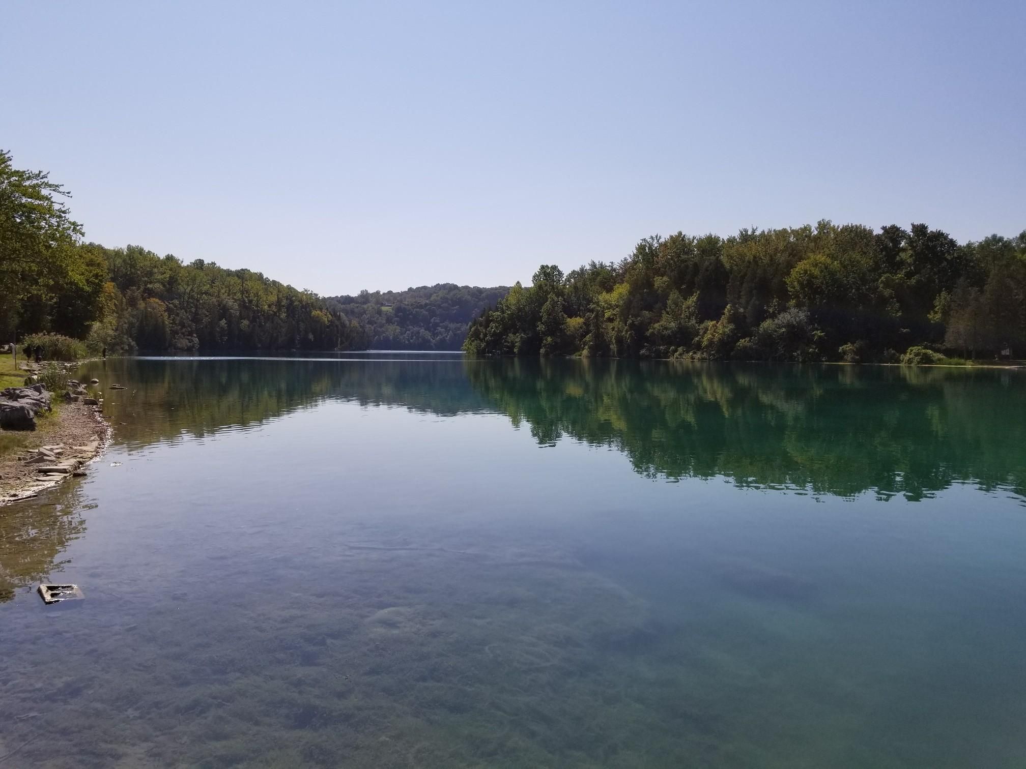 Calm blue-green water reflects green trees under a clear, sunny sky. - Green Lake State Park loop from Fayetteville-Manlius High School