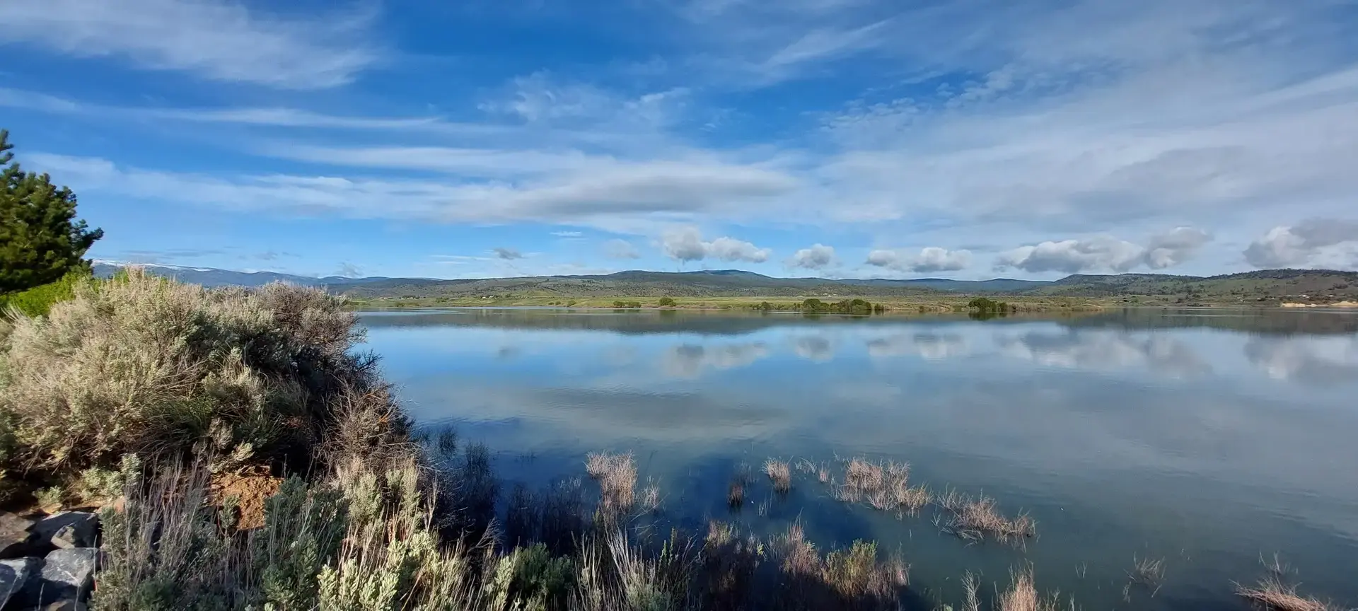 Calm lake reflecting a partly cloudy sky with distant hills and foreground brush.