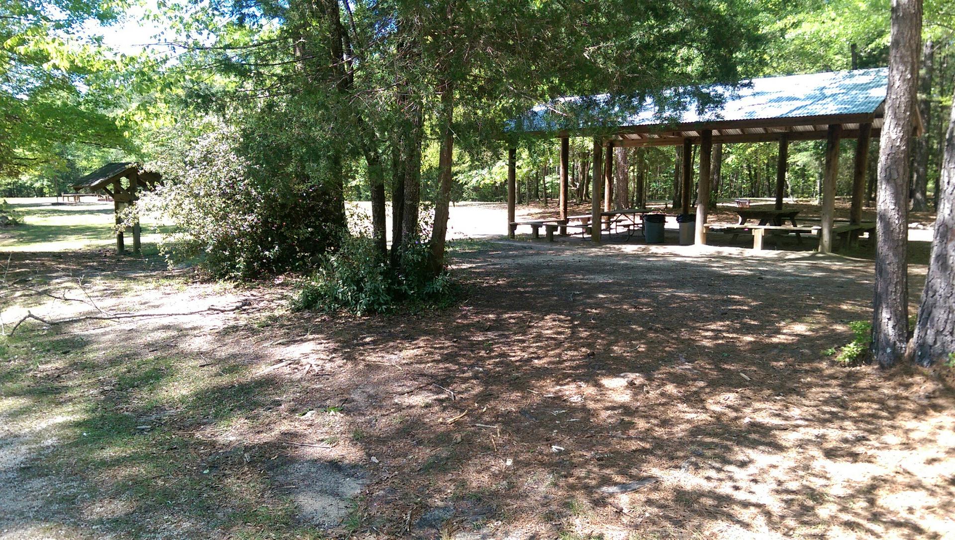 A large wooden picnic shelter in a sunny, wooded park area with dappled light.