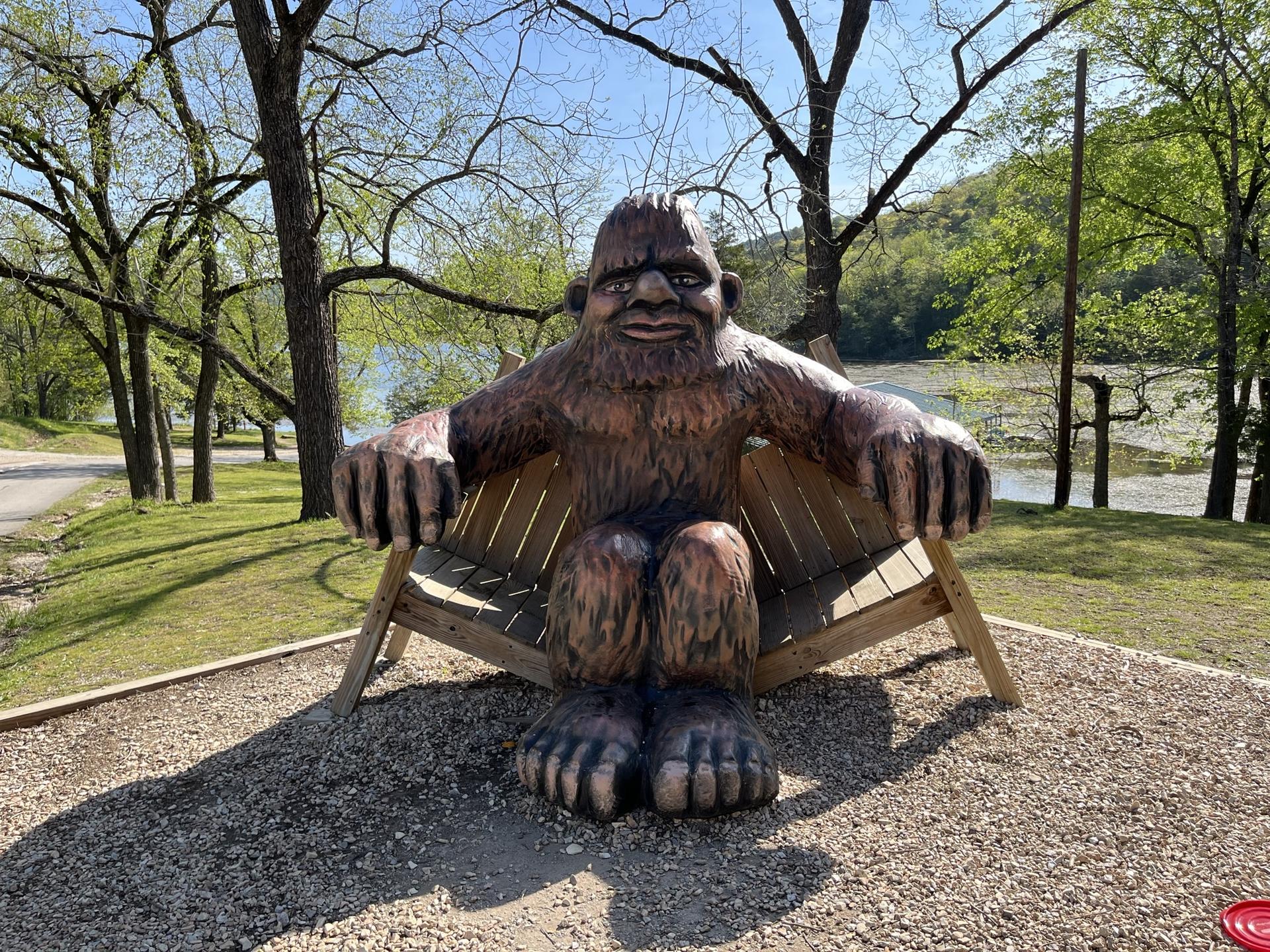 Large Bigfoot statue sitting on a wooden bench in a sunny outdoor setting.
