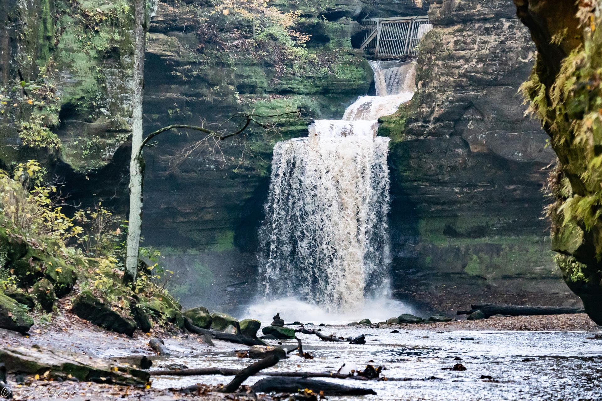 Powerful waterfall plunges down mossy rock cliffs into a stream with fallen logs.