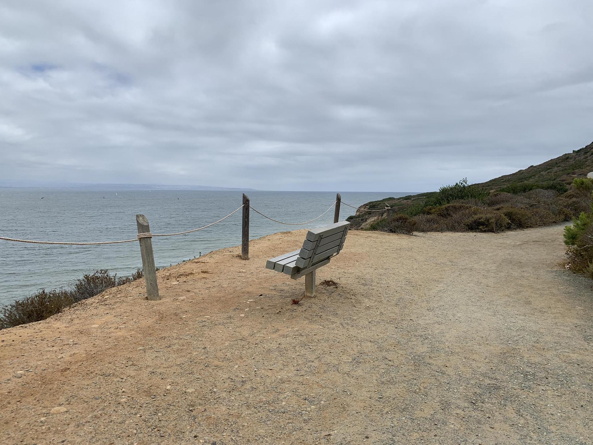 Wooden bench on a dirt path overlooking a calm ocean under cloudy skies.