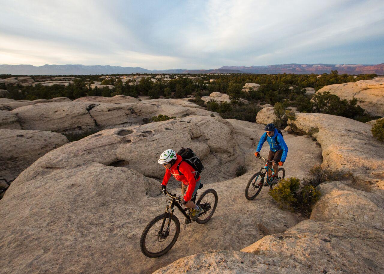 Two mountain bikers ride across rolling rock formations at golden hour.