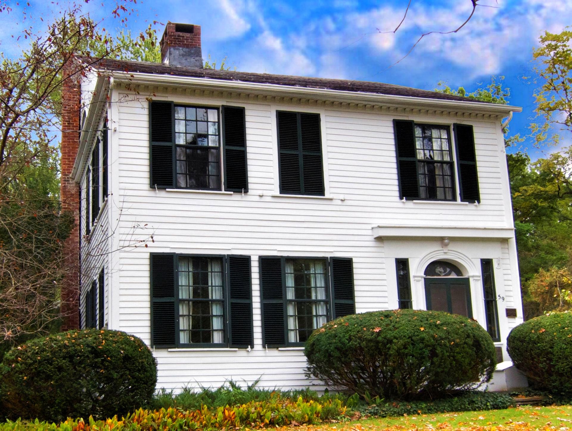 White two-story house with black shutters and a brick chimney, surrounded by bushes.