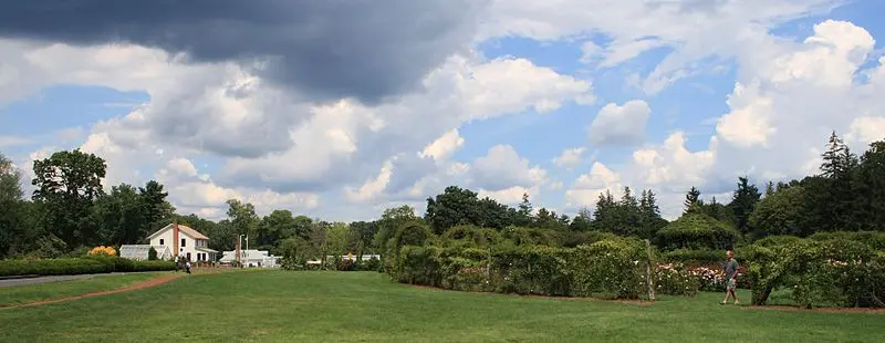 Green field, white building, and hedges under a bright, cloudy sky, with people.