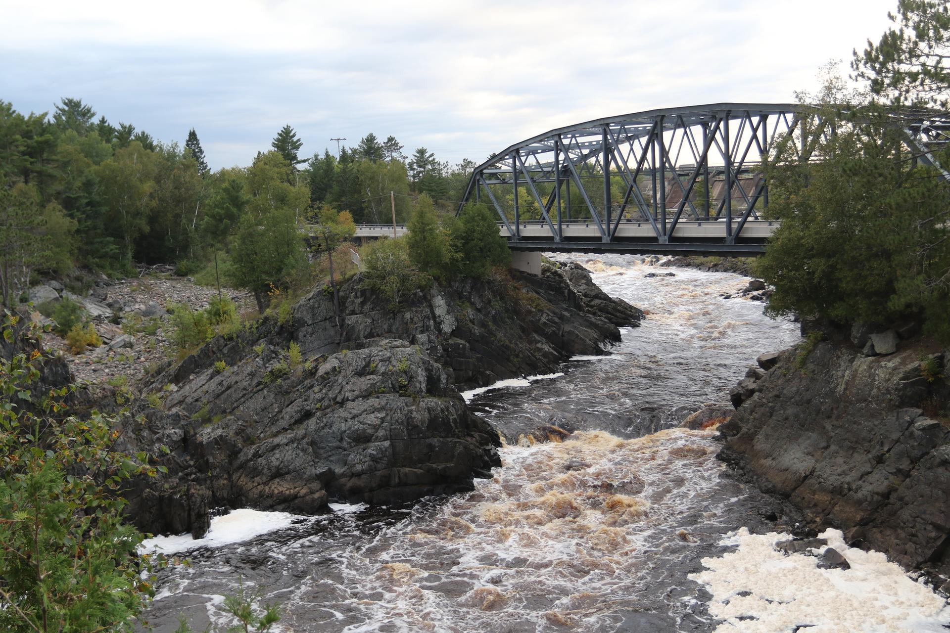 Metal truss bridge over a foamy, brown river with rocky banks and trees.