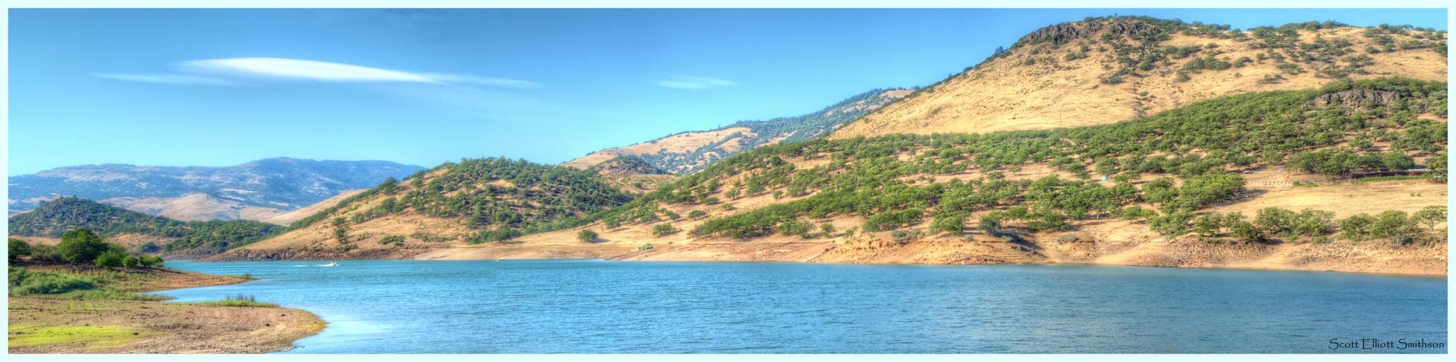 Bright blue lake with dry, forested hills under a clear sky.