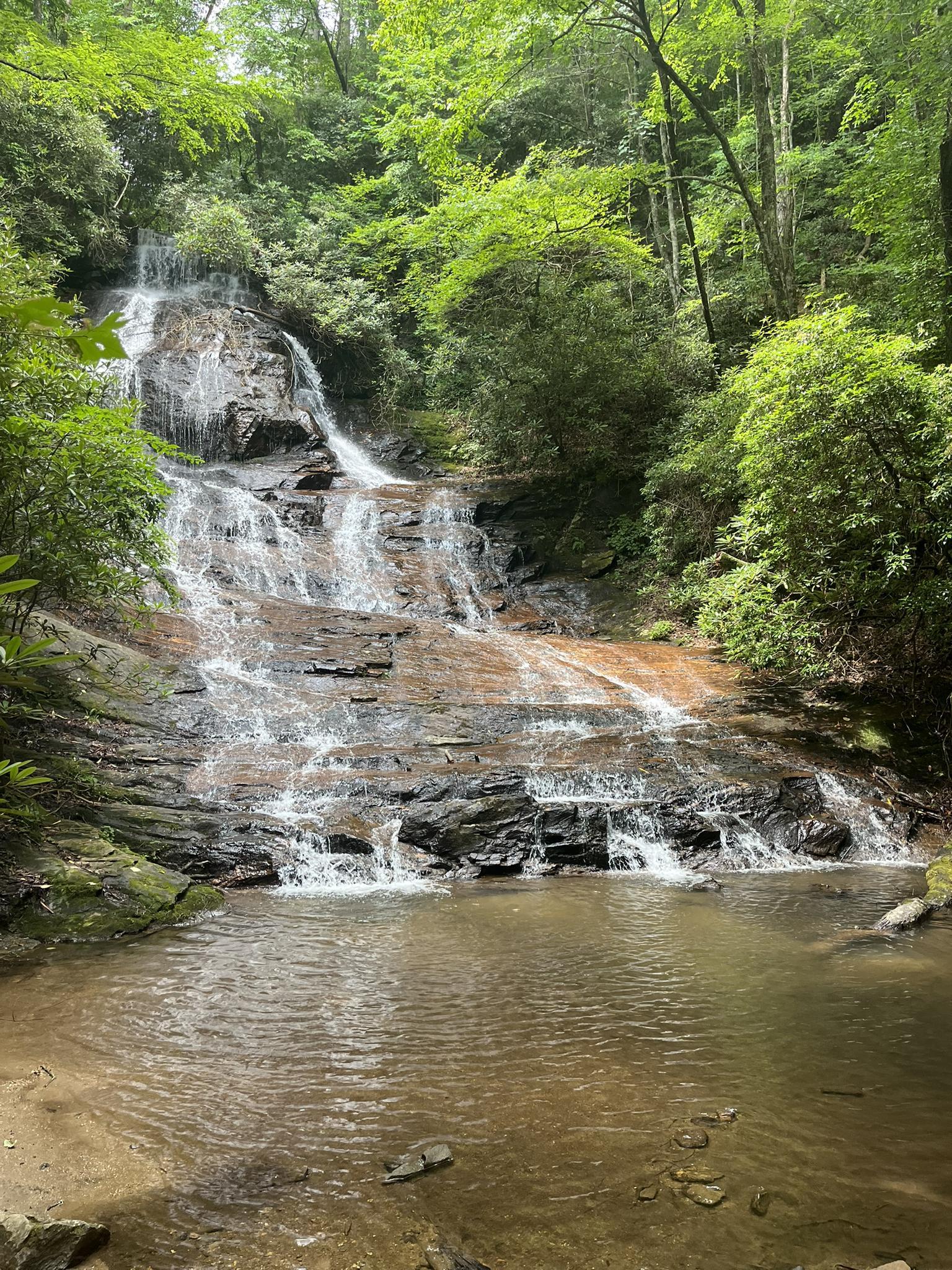 Tiered waterfall flows over wet rocks into a calm pool, framed by dense green trees.