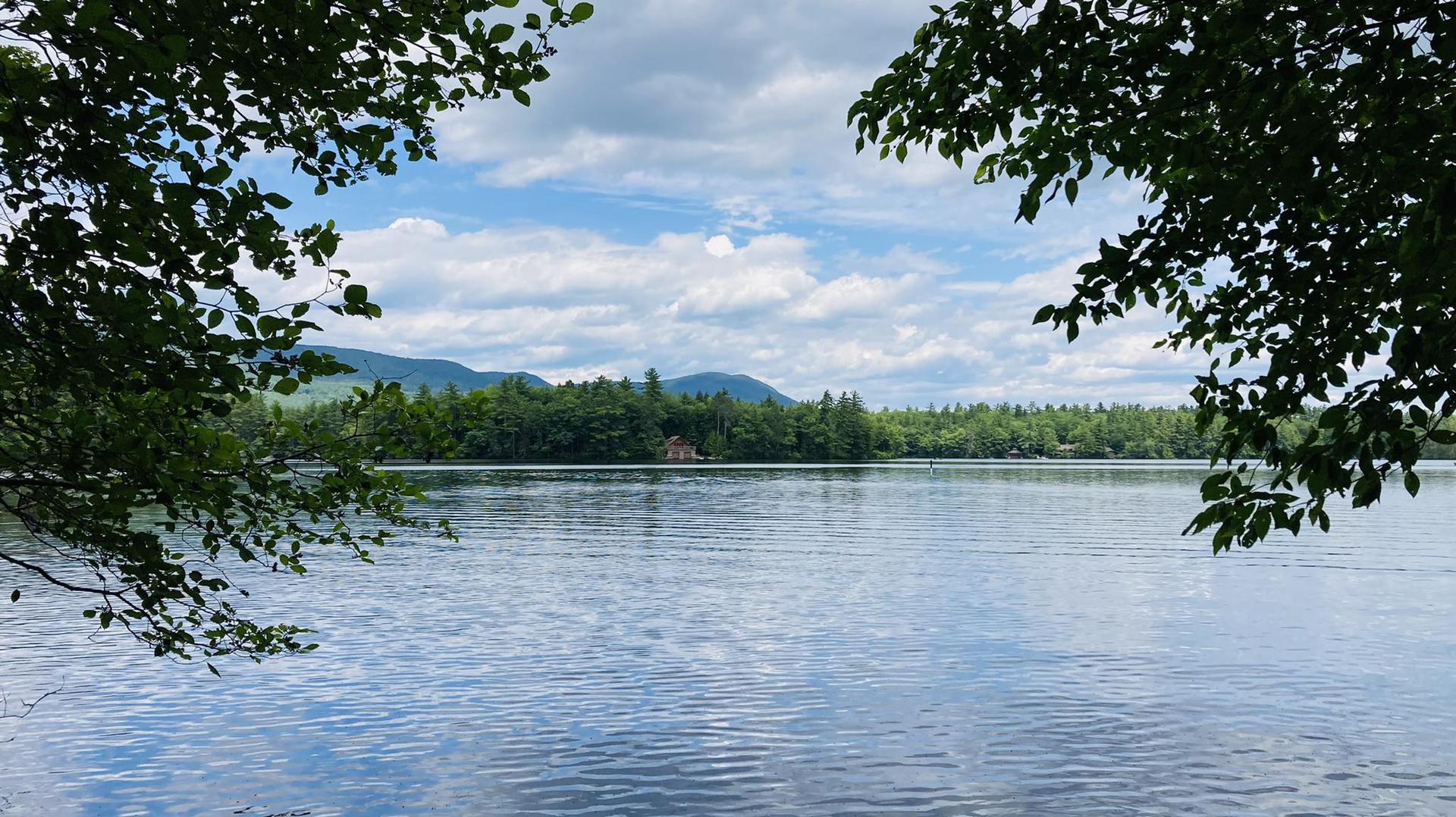 Calm lake framed by trees, with distant forested mountains under cloudy sky.