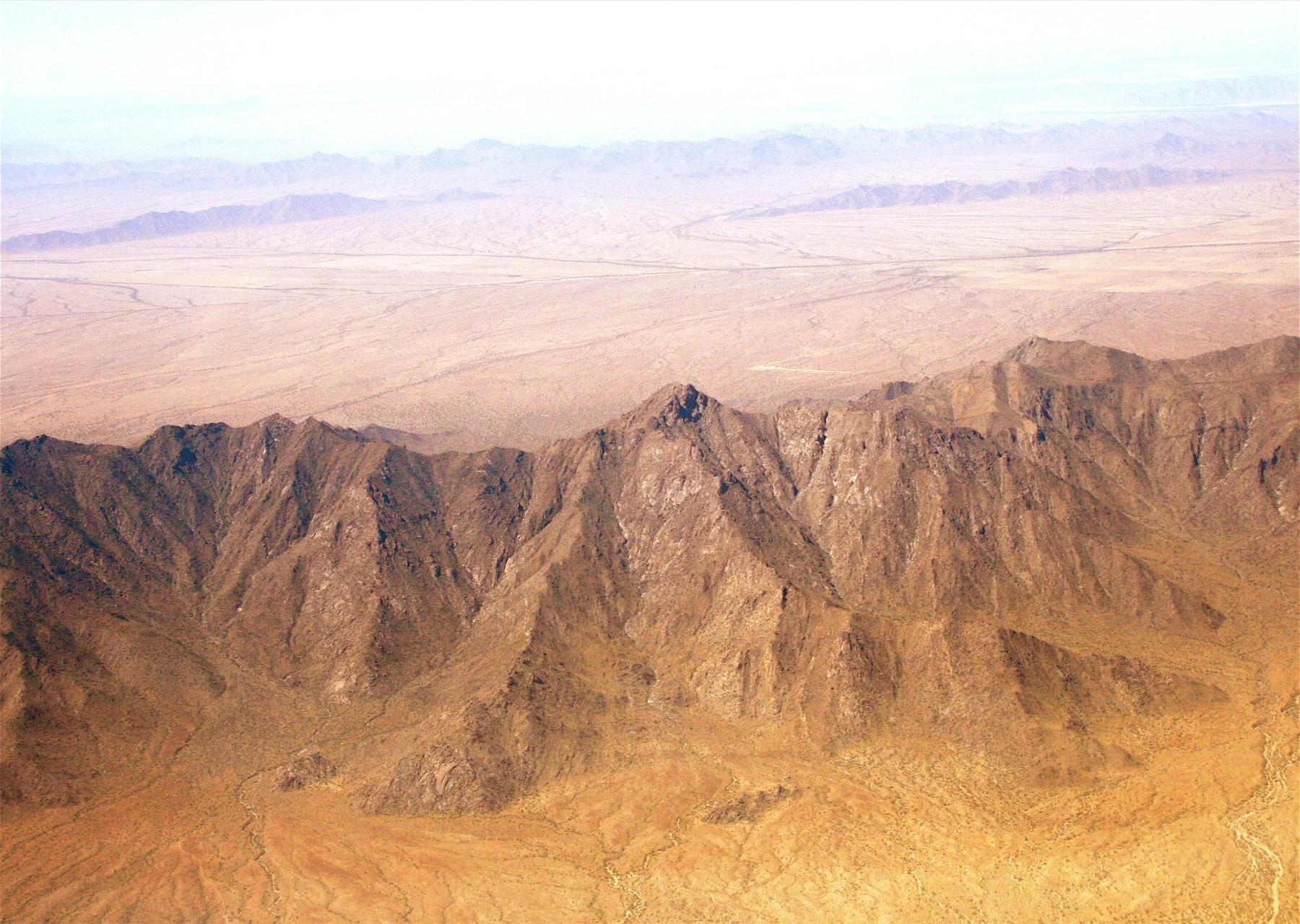 Aerial view of rugged, sunlit desert mountains overlooking a vast, flat plain.