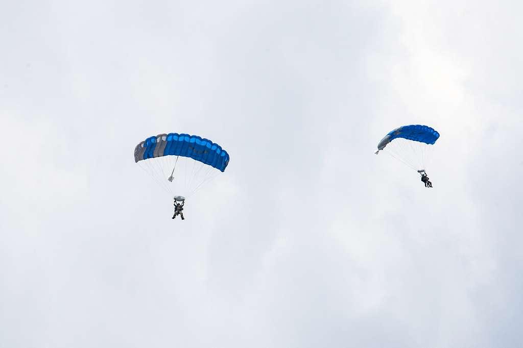 Two skydivers with blue parachutes descend against a bright, cloudy sky.