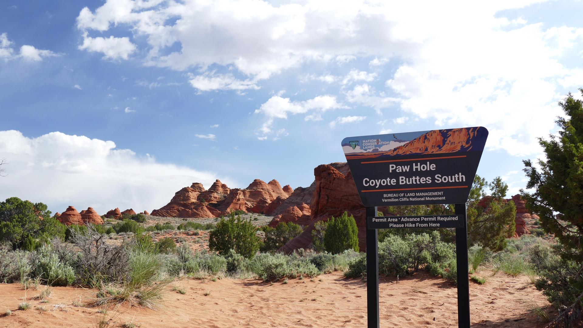 A dark information sign stands in a sandy desert with distant rock formations.