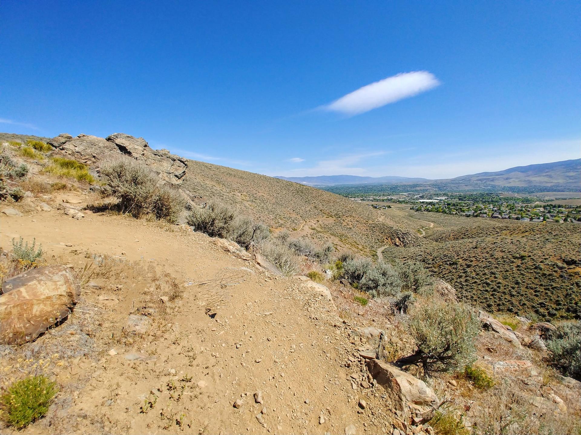 Sandy trail on a steep, brushy hillside overlooking a valley under a clear blue sky.