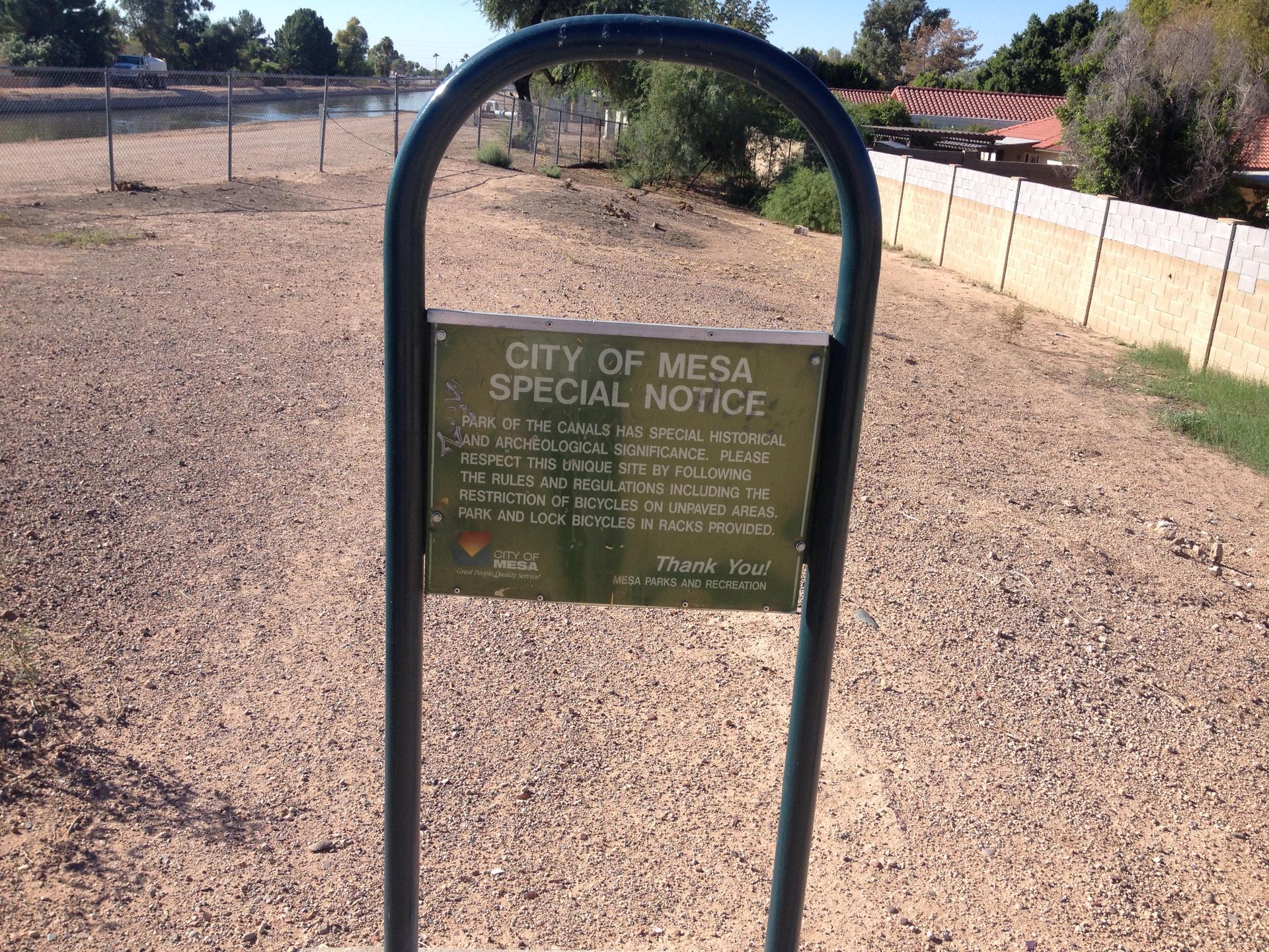 A green notice sign stands on dry, dusty ground beside a canal.