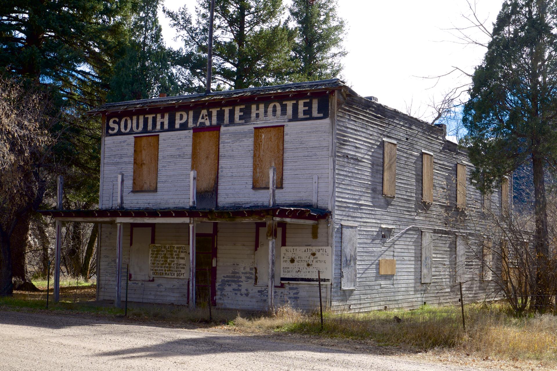 Weathered wooden hotel building with boarded windows, surrounded by trees in daylight.