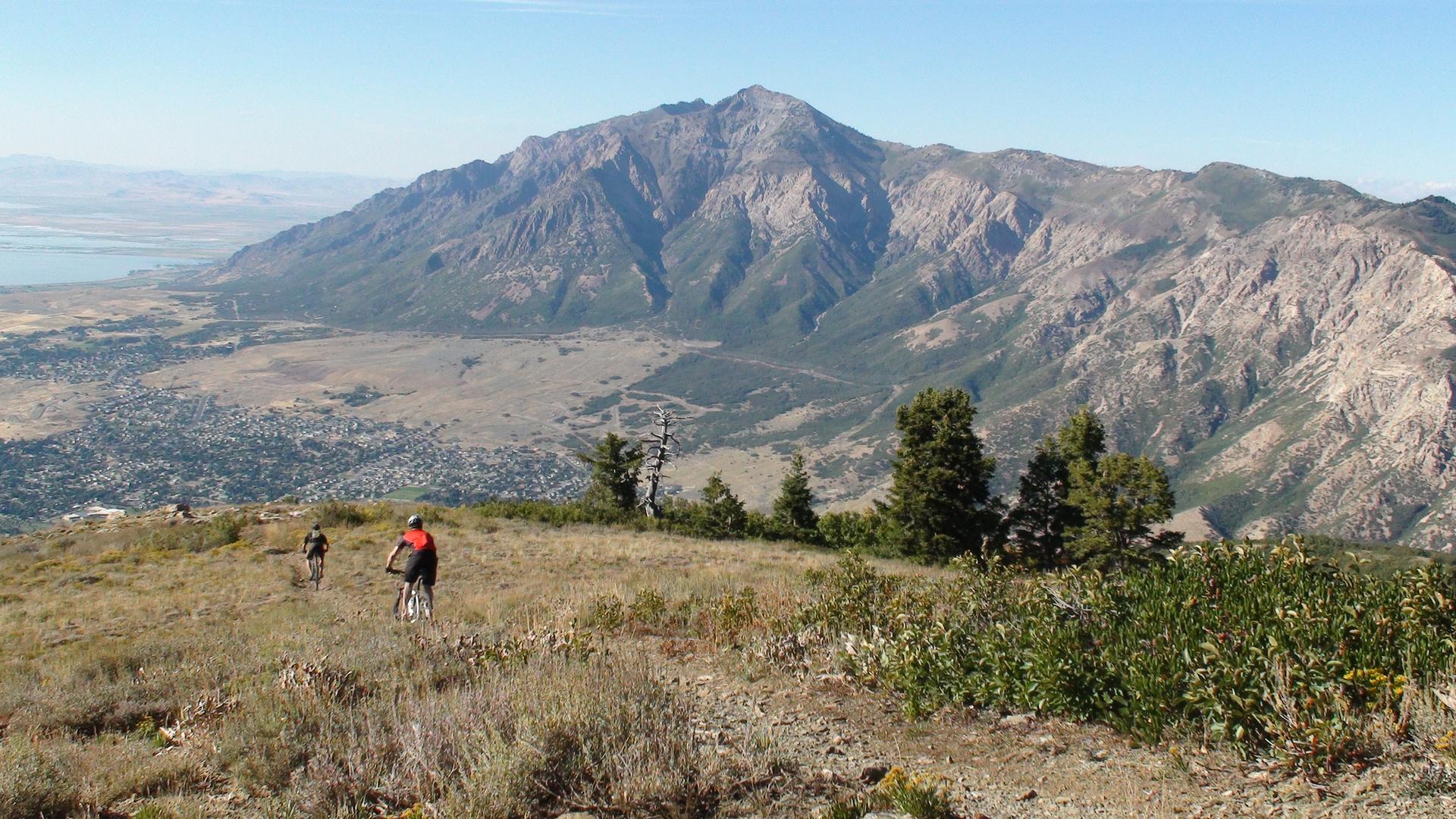 Two mountain bikers on a sunny mountain ridge overlooking a vast valley.