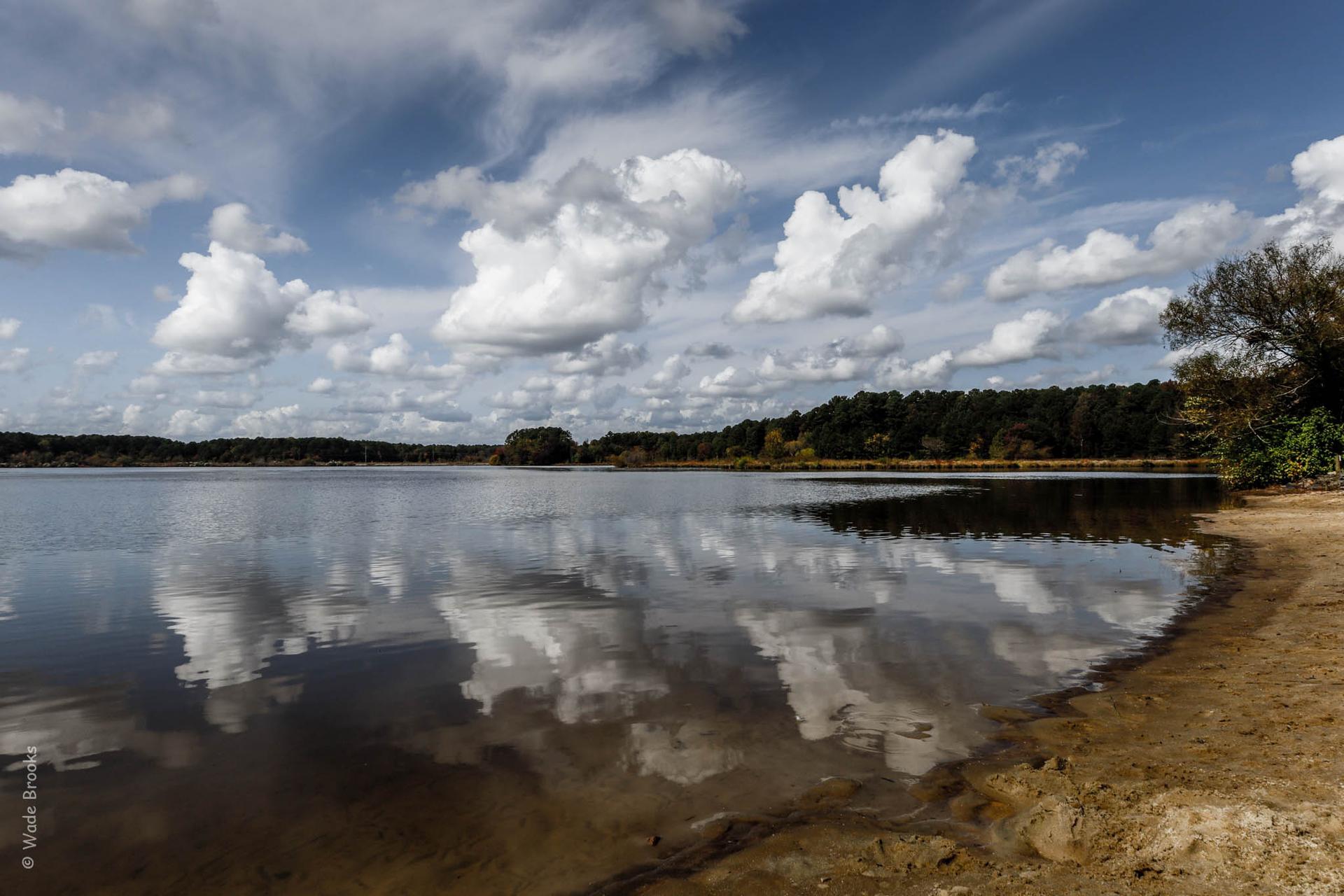 Lake with sandy shore, reflecting dramatic clouds and distant trees.