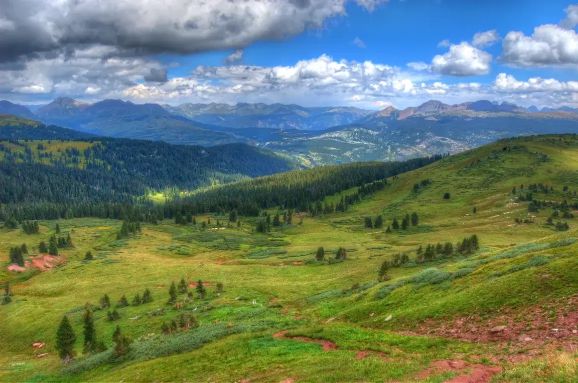 Vast mountain landscape with green hills, forested valleys, and distant peaks under a cloudy sky.