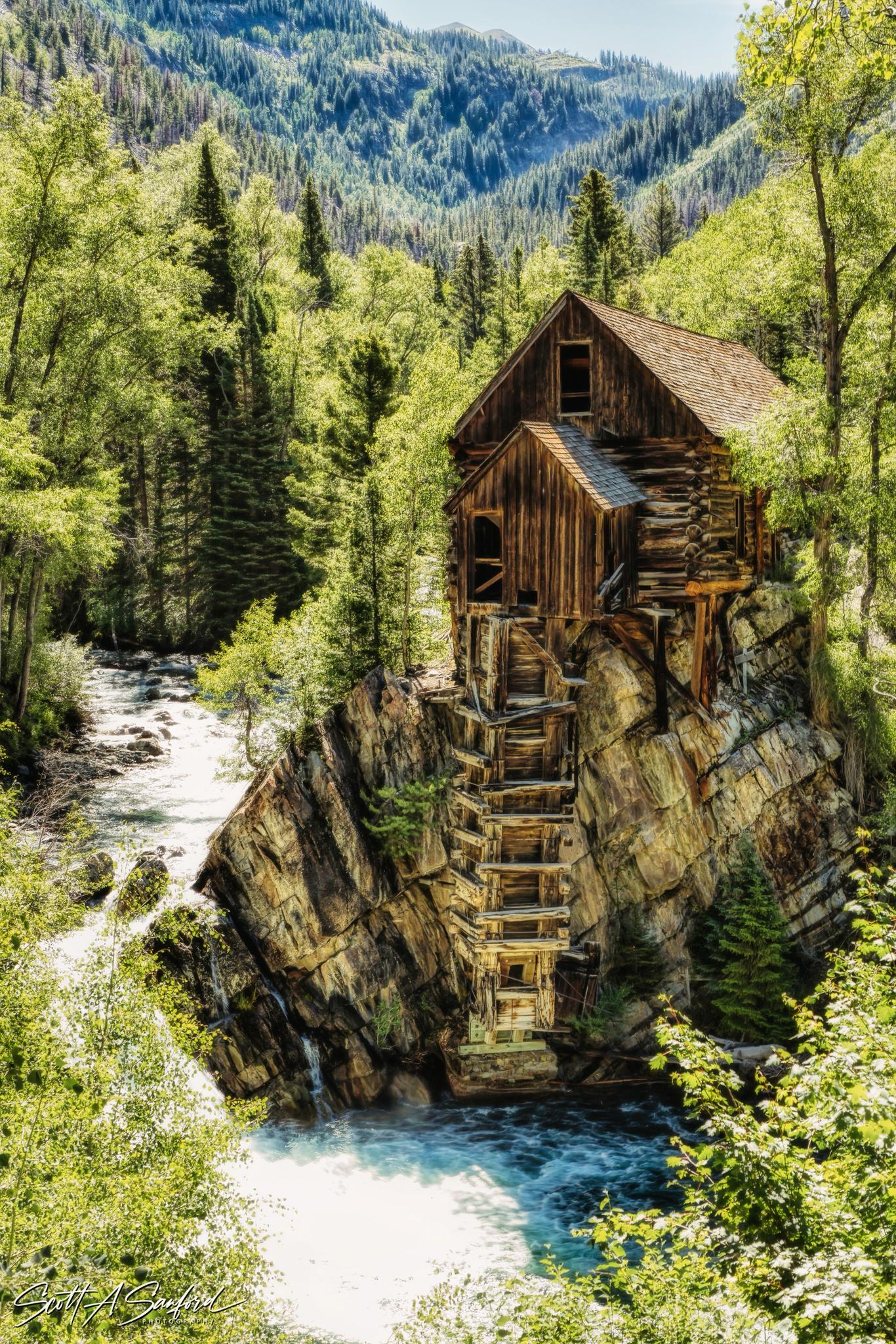 Historic wooden mill building perched on a rock beside a rushing river in a green forest.