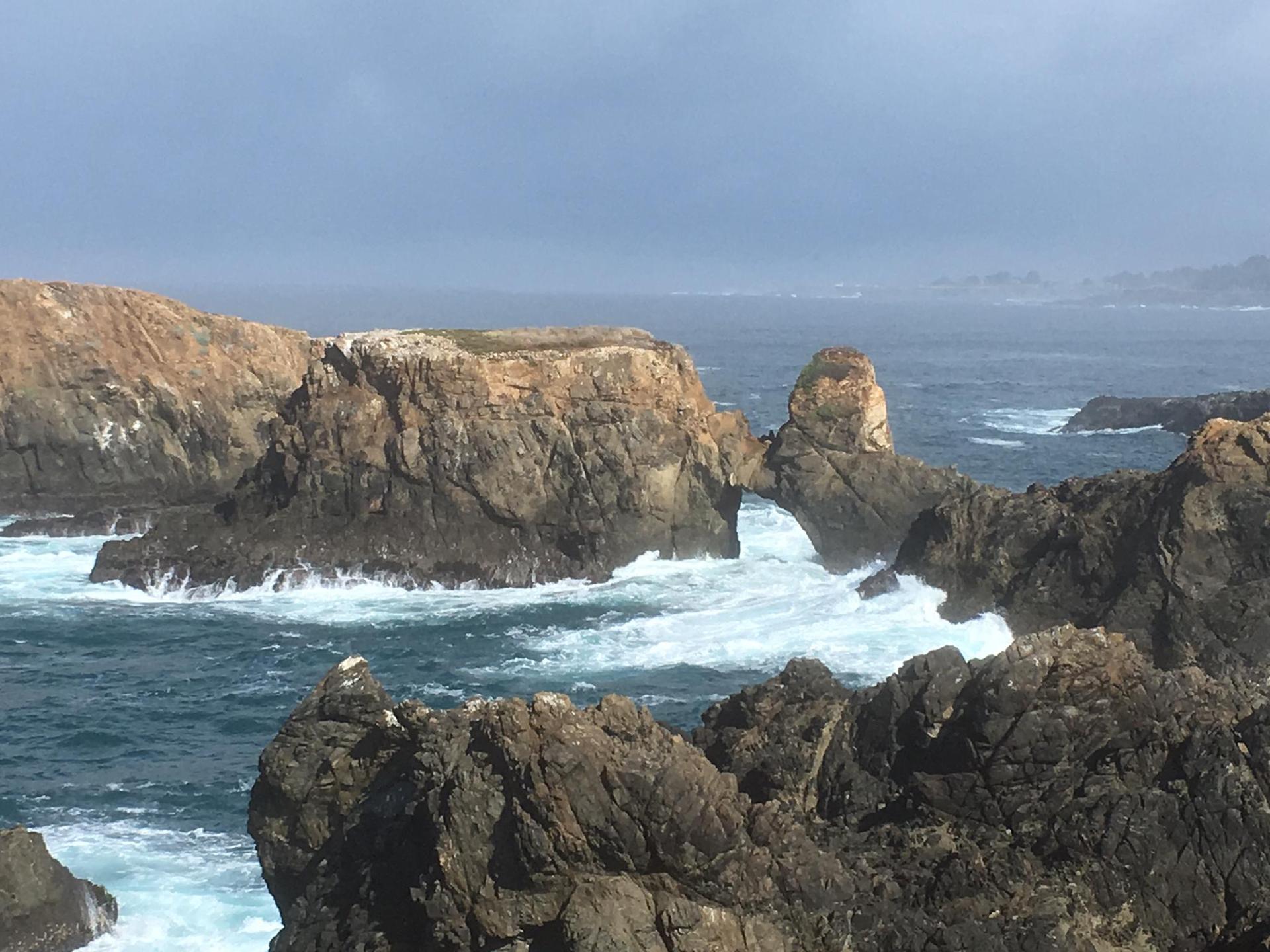 Rough ocean waves crash against rocky sea arches under a cloudy sky.
