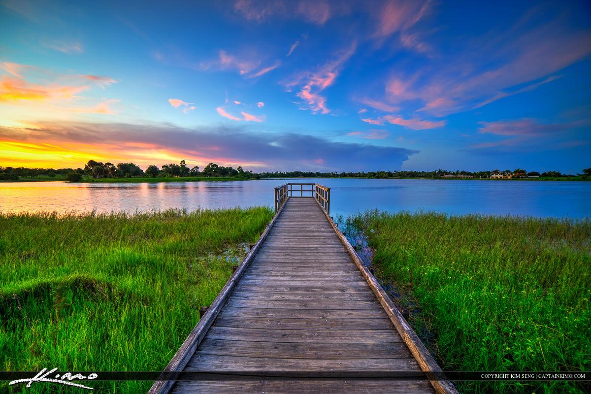 Serene sunset over a lake with a wooden pier and green marsh grass.