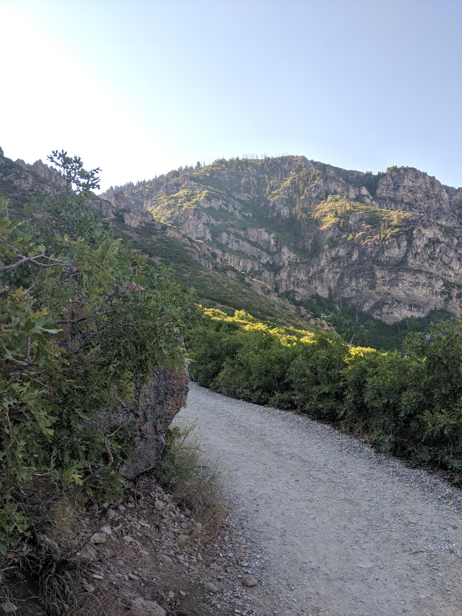 A winding dirt path leads towards a large, sunlit mountain range.