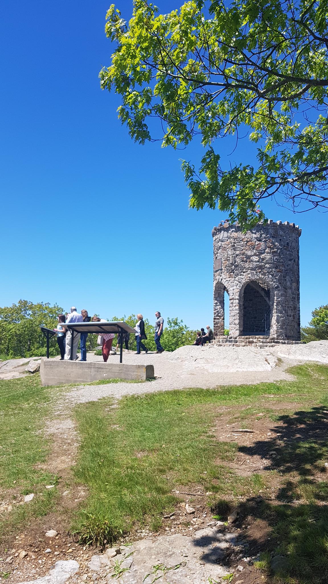 People gather near a stone tower on a sunny, grassy hilltop.