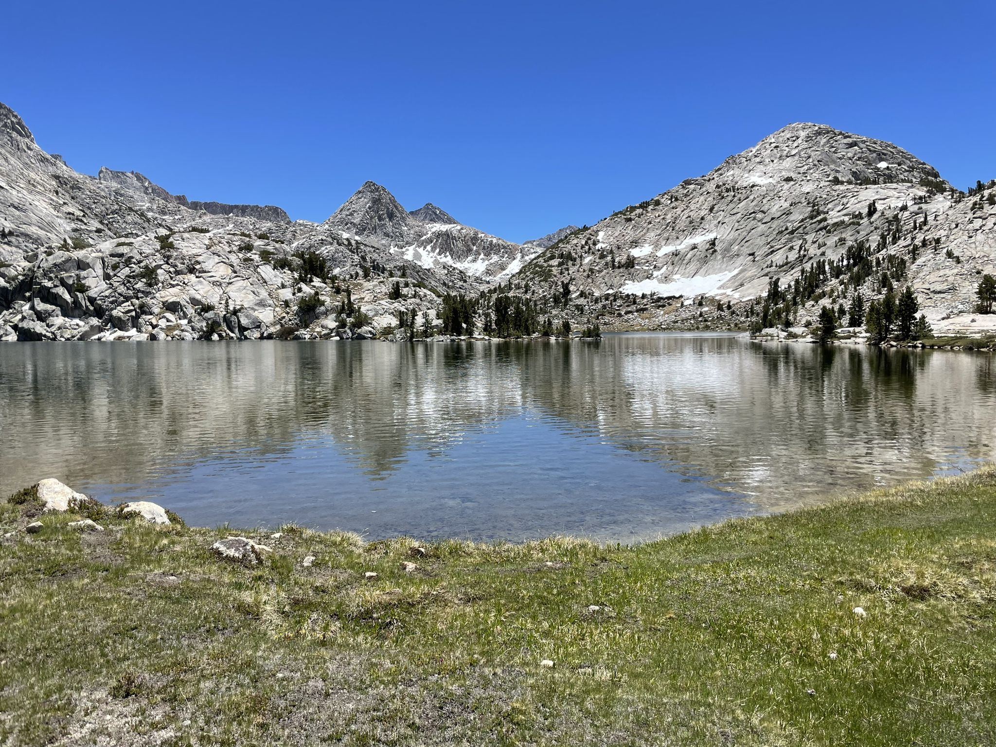 A calm mountain lake reflects rocky peaks and sparse trees under a clear sky. - Florence Lake to Evolution Valley Trail