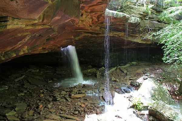 Two waterfalls cascade from a dark rock overhang into a rocky stream.
