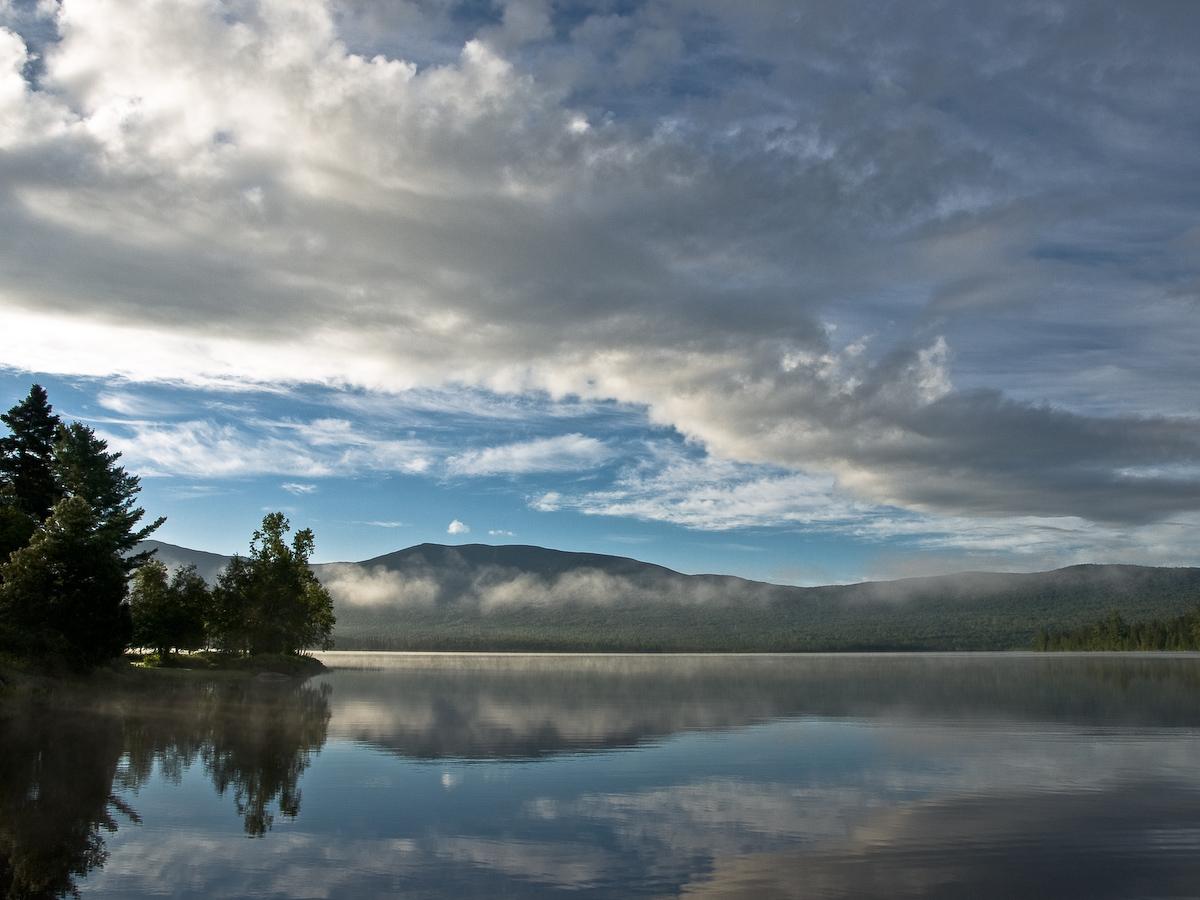 Calm lake reflecting cloudy sky and misty mountains at sunrise.