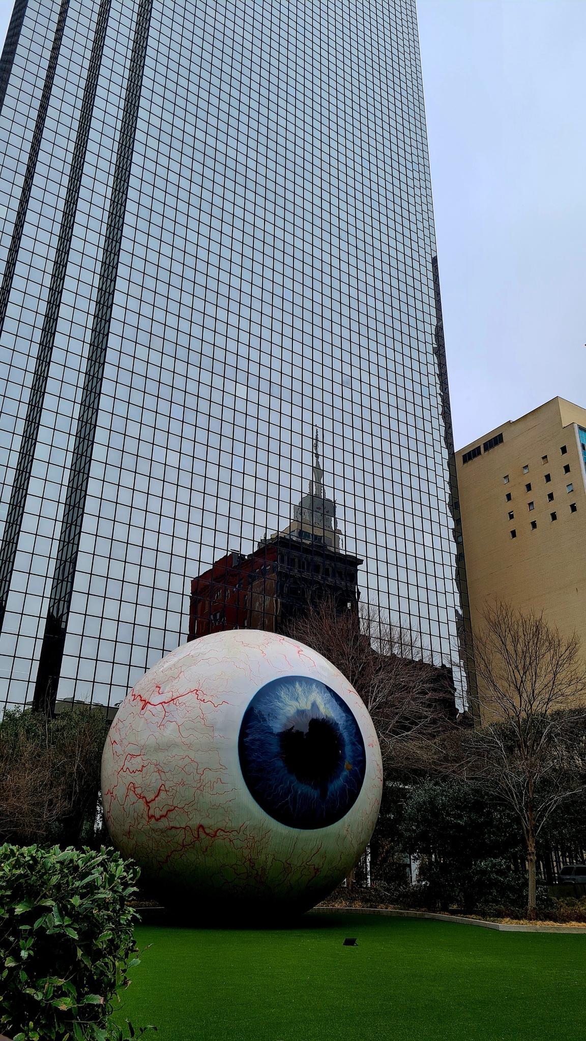 A giant eyeball sculpture sits on grass in front of a tall, modern building.