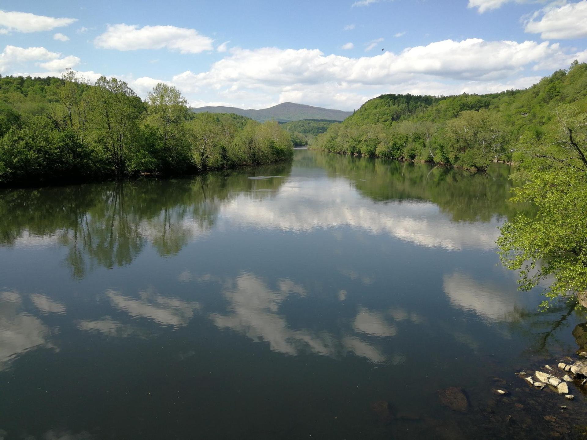 Wide river reflecting blue sky and white clouds, flanked by green forested hills.